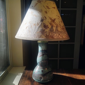 May include: A ceramic table lamp with a teal and brown base and a beige lampshade. The lampshade features a leaf pattern. The lamp sits on a wooden table near a window.