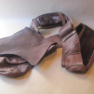 May include: A brown leather shoulder bag with a textured finish. The bag features a wide strap and a flap closure. The leather has a rich, dark brown color, and the bag appears to be handmade.