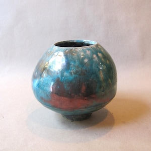 May include: A round ceramic vase with a glossy finish featuring a blend of vibrant turquoise, deep blue, and metallic copper tones. The surface displays a crackled texture with subtle variations in color intensity across the spherical body.