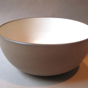 May include: A ceramic bowl with a beige exterior and a white interior. The bowl has a smooth, rounded shape and a simple, minimalist design. The rim of the bowl is a darker shade of beige. This bowl is suitable for serving food.