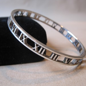 May include: A silver bangle bracelet with Roman numeral markings around the entire circumference.