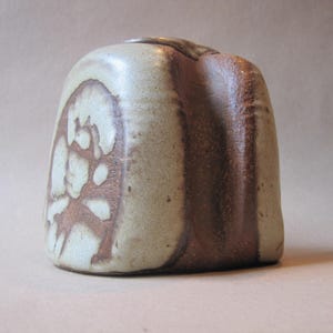May include: A handmade ceramic vase with a unique, organic shape. The vase features a light beige base with brown, abstract patterns on one side and a vertical brown stripe on the other. The vase is approximately 6 inches tall.