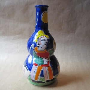 May include: A blue ceramic vase with a double gourd shape. The vase is decorated with a colorful painting of a person wearing a yellow hat and holding a yellow object. The background is a dark blue with yellow and white accents.