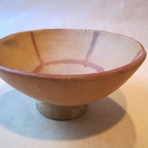 May include: A shallow, tan-colored ceramic bowl with a pedestal base. The interior of the bowl features a painted design consisting of a central circle connected to the rim by four evenly spaced vertical lines in a dark reddish-brown pigment.