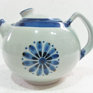May include: A ceramic teapot with a rounded body, a curved handle, and a spout. The teapot is light gray with blue accents and a blue floral design on the front. The lid is also light gray with a blue band.