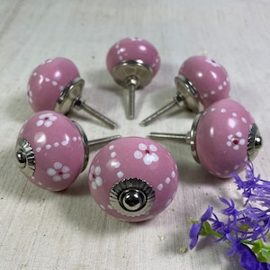 May include: Six pink ceramic cabinet knobs with white floral designs and silver metal bases.