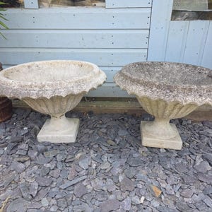 May include: Two weathered stone urns with scalloped rims and square bases. The urns are light grey and sit on a bed of grey stones. A rusty metal planter with a plant is visible on the left.
