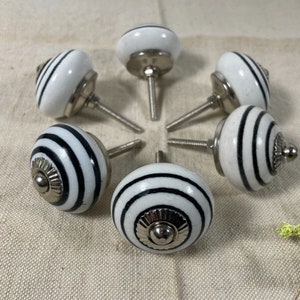 May include: Set of six white ceramic cabinet knobs with black stripes and silver accents. The knobs are round and designed for use on drawers or cabinets.