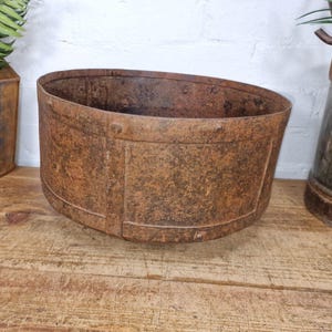 May include: A rusty, oval-shaped metal planter with a riveted construction and a brown finish. The planter has a slightly sloped design. It is displayed on a wooden surface, suggesting a rustic or vintage aesthetic. The planter is empty.