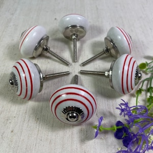 May include: Six white ceramic cabinet knobs with red stripes and silver metal screws. The knobs have a slightly raised, rounded design.