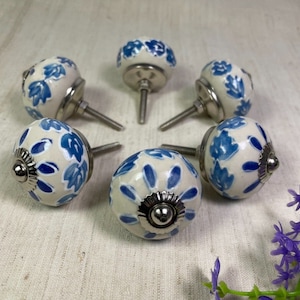 May include: Six white ceramic cabinet knobs with blue floral designs and silver accents. The knobs are round and have a screw-on design.
