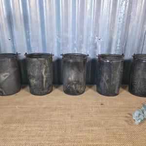 May include: Five aged metal buckets with small handles. The buckets are cylindrical with a dark, weathered appearance, and are arranged in a row. The background features a corrugated metal wall and a hessian surface.
