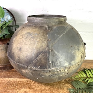 May include: A large, round, metal planter with a distressed, aged finish. The planter has a wide opening at the top and is made of galvanised metal. It has a rustic, industrial look.