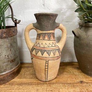 May include: A terracotta vase with two handles and a geometric pattern in black, red and white. The vase is sitting on a wooden surface.