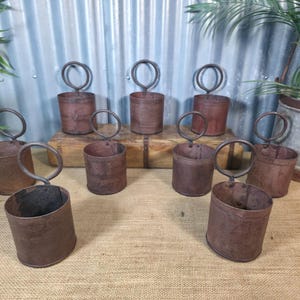 May include: Ten rustic metal buckets with circular handles. The cylindrical buckets have a weathered, brown finish, giving a vintage look. They are arranged on a burlap surface with a corrugated metal backdrop, suggesting an antique aesthetic.