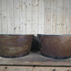 May include: Two large, oval-shaped metal wash tubs with a weathered, rusted brown patina. Each tub features a wide, reinforced rim and a dark metal band around the base. The right tub includes a circular metal handle attached to the side.