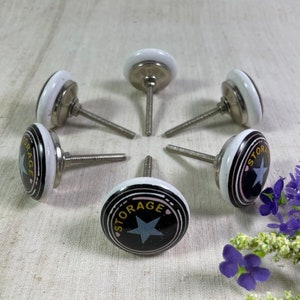 May include: Six white ceramic knobs with black and gold accents. Each knob features a star and the word "STORAGE" in a circular design. The knobs have silver metal stems.