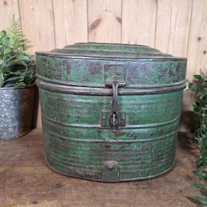 May include: A round, weathered green metal storage container with a domed lid and a central vertical metal latch mechanism. The surface shows signs of oxidation and paint wear, revealing dark brown metal underneath the chipped green finish.