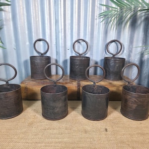 May include: Seven aged metal planters with circular handles. The cylindrical planters have a dark, weathered finish. They are arranged on a wooden box and burlap surface. The background features corrugated metal and greenery.