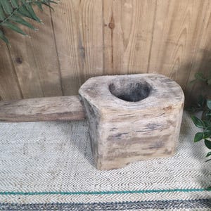May include: A weathered wooden mortar and pestle. The mortar is a large, square block with a central bowl and a long, rectangular handle. The wood has a natural, aged appearance, with visible grain and knots. The pestle is a simple, cylindrical piece of wood.