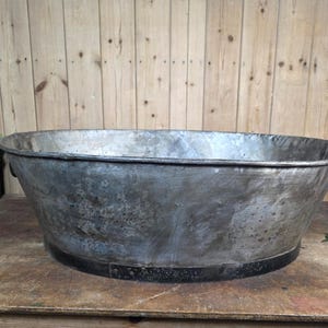 May include: A large, oval-shaped galvanised metal wash tub with a weathered, mottled grey finish. The basin features a wide, reinforced dark metal band around the base and small, curved metal handles attached to the sides for lifting and transport.