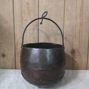May include: A dark, weathered metal bucket with a rounded base and a curved handle featuring a central loop. The exterior surface displays visible horizontal bands secured with small rivets, showing a textured, aged patina across the entire body.