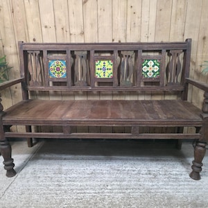 May include: A dark brown wooden bench featuring a high backrest with vertical slats and three square decorative tiles. The tiles display colourful floral patterns in shades of yellow, green, blue, and red. The bench has turned legs and armrests.