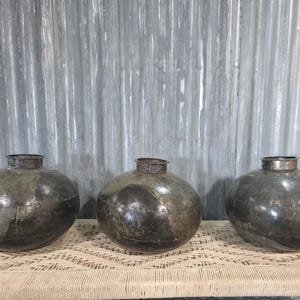May include: Three antique, dark metal, round vessels with a wide body and narrow neck. The vessels are displayed on a woven surface against a corrugated metal background. The vessels have a rustic, aged appearance.