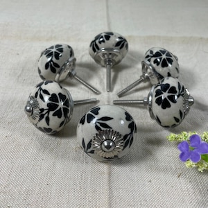 May include: Six white ceramic knobs with black floral designs and silver metal accents. The knobs are round and arranged in a circle on a white fabric background.