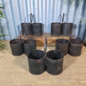 May include: Eight dark metal containers with handles, showing a rustic, aged appearance. The cylindrical containers have circular handles attached to the sides. They are arranged on a burlap surface, with a corrugated metal background.