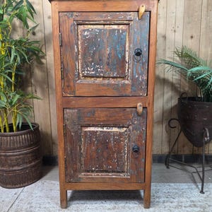 May include: A tall, rectangular wooden cabinet featuring two stacked doors with a distressed, weathered finish in shades of brown and blue. Each door is equipped with a small, dark metal ring pull handle and a simple wooden latch mechanism.