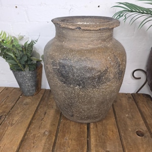 May include: A large, weathered, brown ceramic vase with a wide opening. The vase has a textured surface and dark patches. It sits on a wooden table with other decorative items, including a metal bucket with greenery and a potted plant.