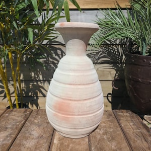 May include: A large, off-white ceramic vase with a wide base and a flared top. The vase has horizontal ridges and a slight reddish-pink hue in areas. It is displayed on a weathered wooden surface, with greenery in the background.