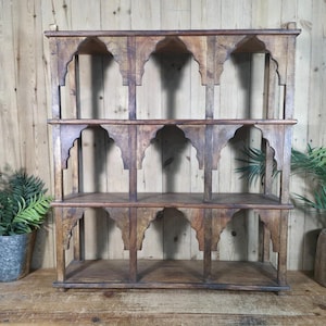 May include: A wooden, three-tiered shelf with an arched design. The shelf is made of dark brown wood and has six open compartments. The shelf is approximately 100 cm wide and 100 cm tall. The shelf is empty.