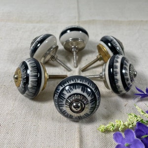 May include: Six black and white ceramic drawer knobs with a silver metal base. The knobs have a striped pattern and are arranged in a circle on a white fabric surface.
