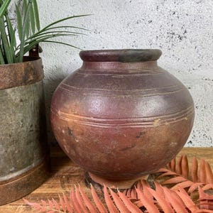 May include: A large, round, terracotta pot with a dark brown glaze. The pot has a slightly textured surface and is decorated with horizontal lines. The pot is sitting on a wooden surface.