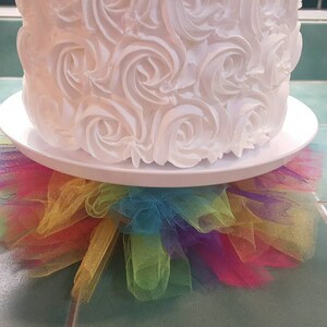 May include: A white cake with a rosette frosting design sits on a white cake stand with a rainbow tulle skirt.