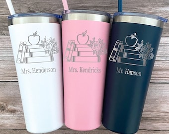 May include: Three stainless steel tumblers with lids and straws. Each tumbler features a design of a stack of books, an apple, and flowers. The tumblers are white, pink, and blue. The tumblers are personalized with the names "Mrs. Henderson", "Mrs. Kendricks", and "Mr. Hanson".