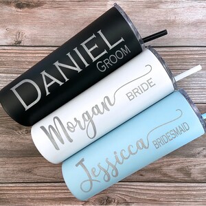 Pode incluir: Três copos de aço inoxidável com tampas e canudos. Os copos são preto, branco e azul claro. O copo preto tem o nome "DANIEL GROOM" gravado. O copo branco tem o nome "Morgan BRIDE" gravado. O copo azul claro tem o nome "Jessica BRIDESMAID" gravado.