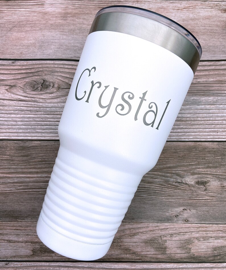 May include: White stainless steel tumbler with a silver lid. The tumbler has a ribbed design and the name "Crystal" is engraved on the front.