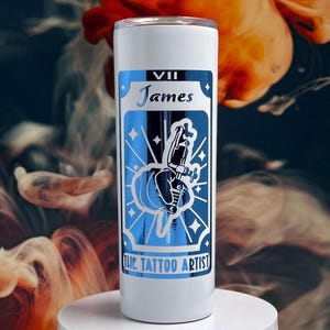 May include: White stainless steel tumbler with a blue and white design. The design features a tattoo machine and the text "VII James The Tattoo Artist".