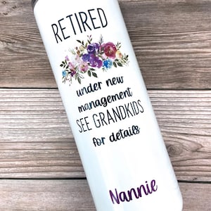Pode incluir: Um copo de aço inoxidável branco com um design floral preto e o texto "Aposentado sob nova gerência veja netos para mais detalhes" e o nome "Nannie" na parte inferior.