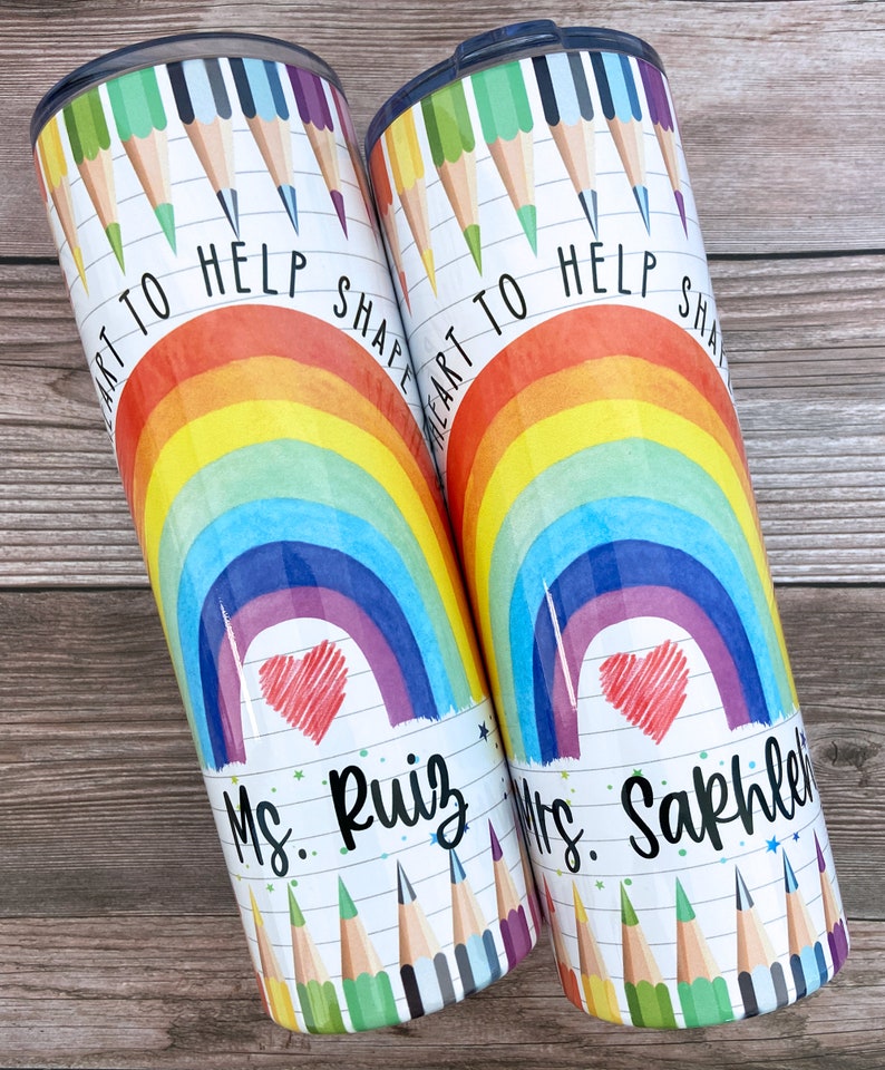 Pode incluir: Dois copos de a&ccedil;o inoxid&aacute;vel com um design de arco-&iacute;ris, cora&ccedil;&atilde;o e l&aacute;pis de cor. O texto "Art to help shape heart" est&aacute; nos copos. Um copo diz "Mrs. Ruiz" e o outro diz "Mrs. Sakhleh".