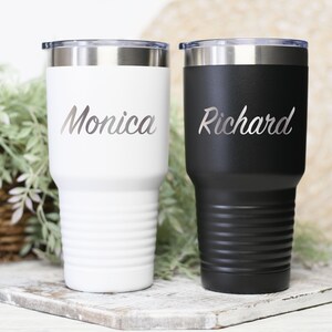 May include: Two stainless steel tumblers with lids. The tumbler on the left is white with the name "Monica" engraved on it. The tumbler on the right is black with the name "Richard" engraved on it.