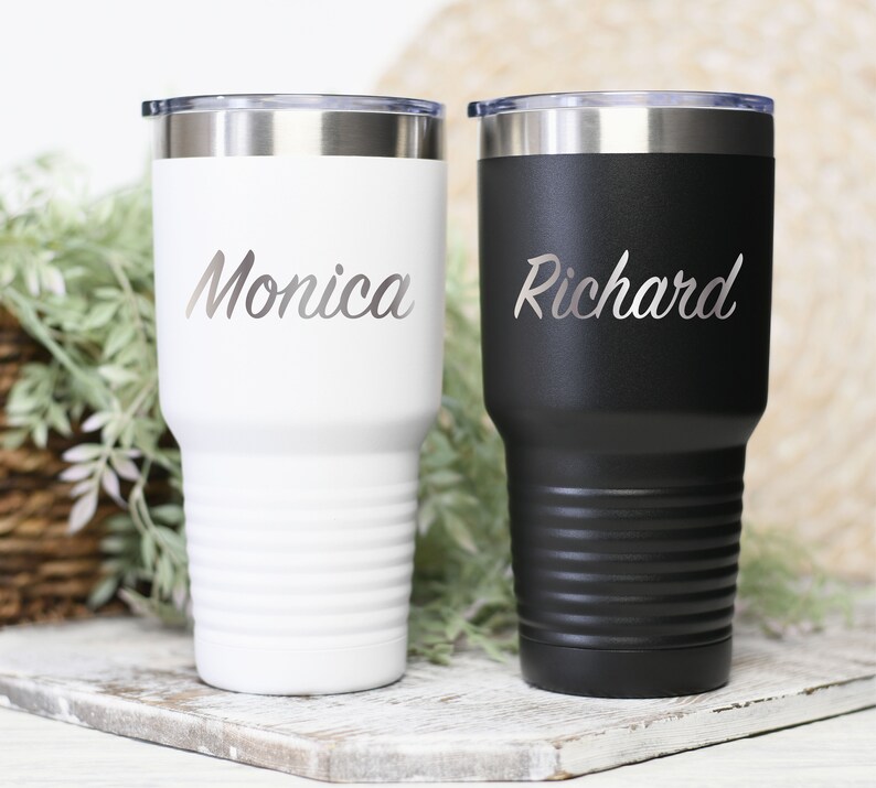 May include: Two stainless steel tumblers with lids. The tumbler on the left is white with the name "Monica" engraved on it. The tumbler on the right is black with the name "Richard" engraved on it.