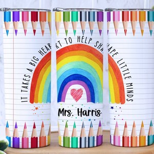 Pode incluir: Tr&ecirc;s copos de a&ccedil;o inoxid&aacute;vel com fundo branco e design de arco-&iacute;ris colorido. Os copos t&ecirc;m um fundo branco com um arco-&iacute;ris, um cora&ccedil;&atilde;o e o texto "&Eacute; preciso um grande cora&ccedil;&atilde;o para ajudar a moldar as mentes pequenas". Os copos tamb&eacute;m t&ecirc;m uma fileira de l&aacute;pis coloridos na parte superior e inferior.