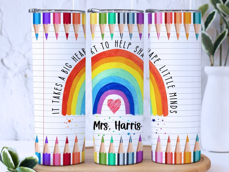 Pode incluir: Tr&ecirc;s copos de a&ccedil;o inoxid&aacute;vel com fundo branco e design de arco-&iacute;ris colorido. Os copos t&ecirc;m um fundo branco com um arco-&iacute;ris, um cora&ccedil;&atilde;o e o texto "&Eacute; preciso um grande cora&ccedil;&atilde;o para ajudar a moldar as mentes pequenas". Os copos tamb&eacute;m t&ecirc;m uma fileira de l&aacute;pis coloridos na parte superior e inferior.