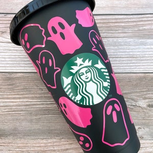 May include: Black reusable cup with a green Starbucks logo and pink ghost silhouettes. The cup has a black lid.