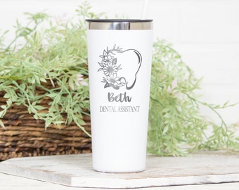 May include: A white stainless steel tumbler with a black lid and straw. The front features a gray graphic of a tooth adorned with flowers, followed by the name Beth and the text DENTAL ASSISTANT printed in a gray sans-serif font.