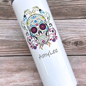 Pode incluir: Um copo branco de aço inoxidável com um design de caveira de açúcar colorido e o nome "Amy Lee" impresso na parte inferior.